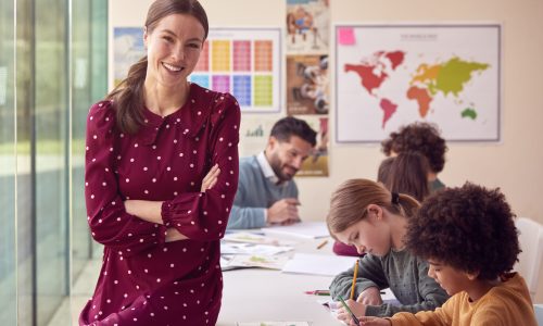 Portrait Of Smiling Female Elementary School Teacher Working At Desk In Classroom With Students